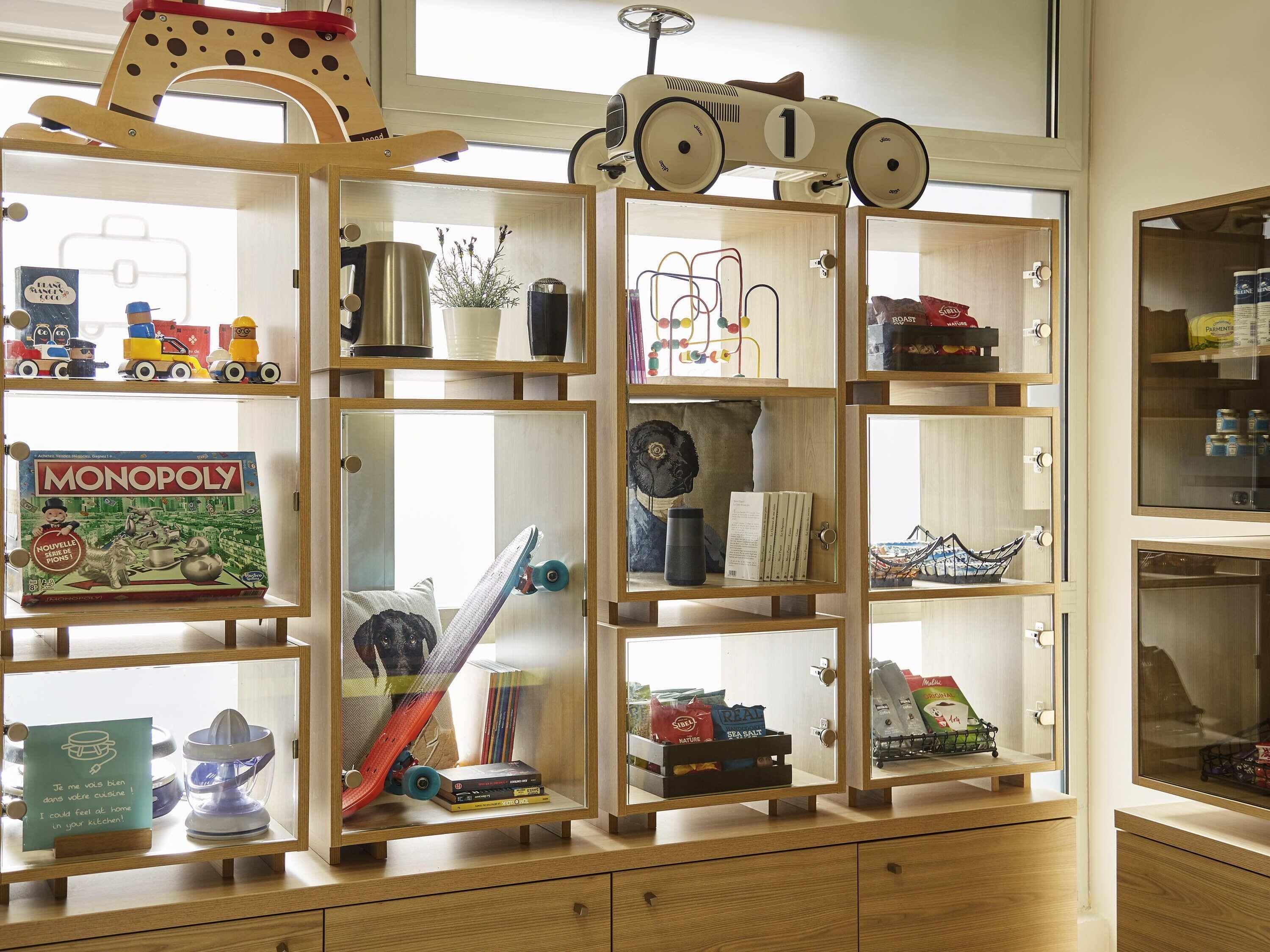 A wooden cabinet with multiple shelves containing various items such as a Monopoly board game, a toy car, and kitchen utensils.