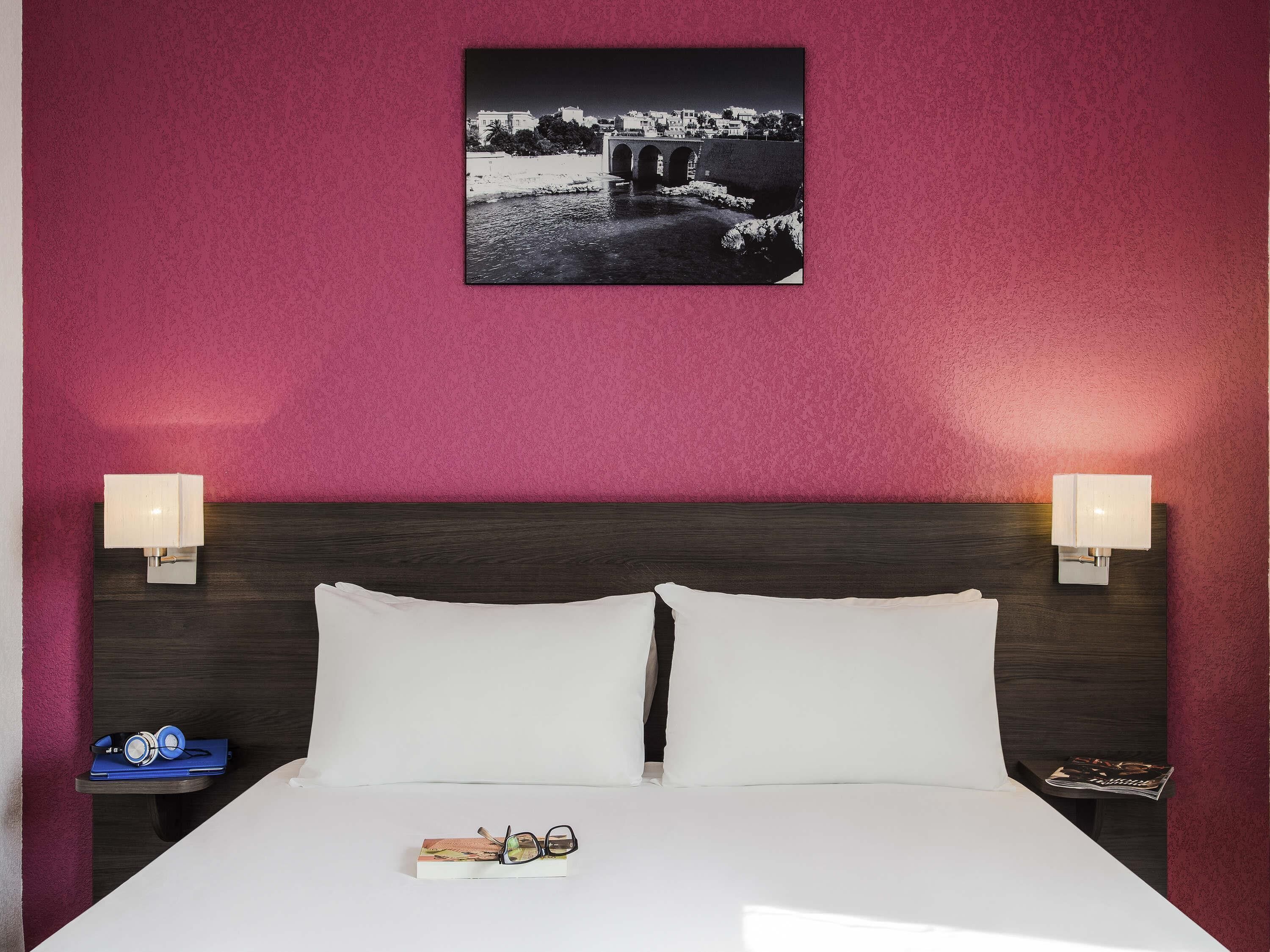 A hotel room with a bed, two bedside lamps, a framed picture on the wall, and a small table with a book and glasses.