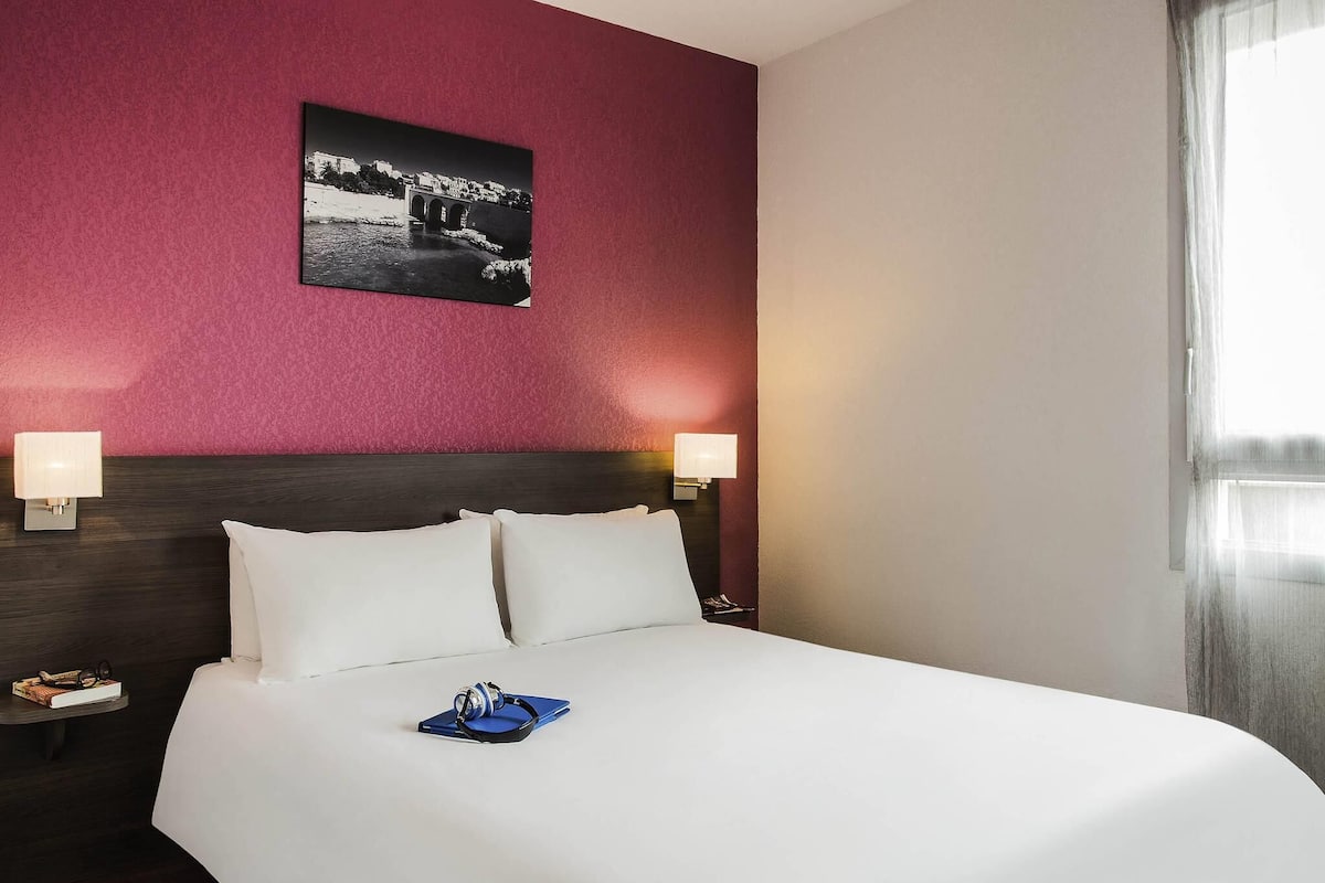 Una habitación de hotel con una cama, dos lámparas de noche, una mesita de noche, un cuadro en la pared y una ventana con cortinas.