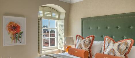 A hotel room with a large bed, a window with shutters, a green upholstered headboard, and a framed artwork on the wall.