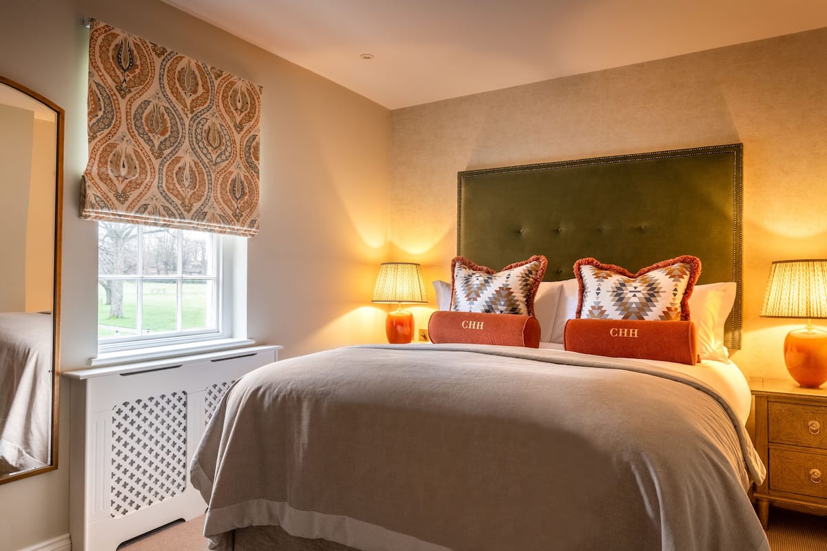 A bedroom with a large bed, two bedside lamps, a headboard, and a window with patterned curtains.