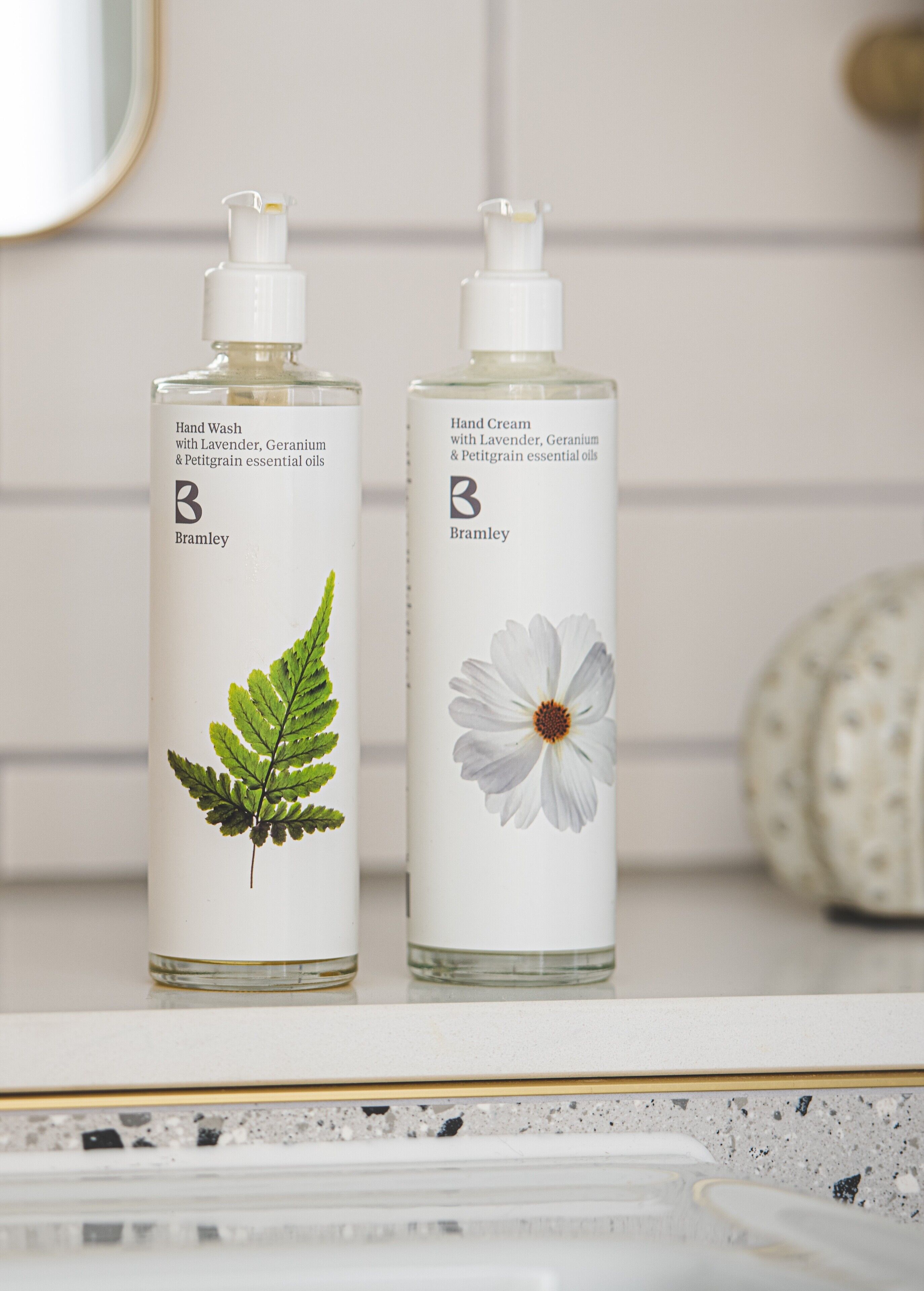 Dos botellas de jabón de manos y crema hidratante con aceites esenciales de lavanda, geranio y petitgrain de Bramley sobre una encimera blanca.