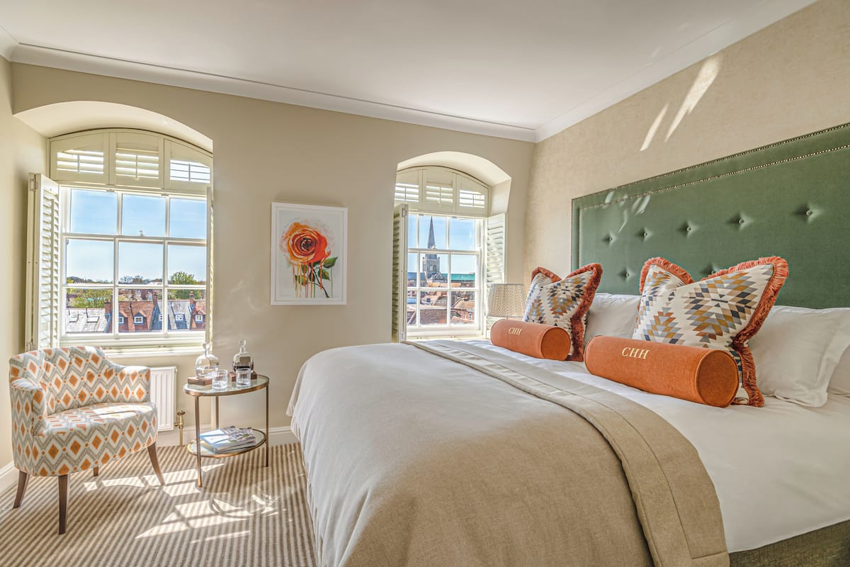 A hotel room with a large bed, two windows, a chair, a small table, and a painting.
