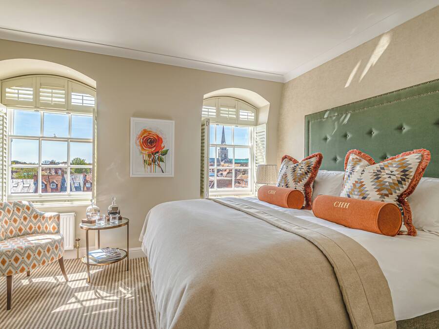 Een hotelkamer met een groot bed, twee ramen, een stoel, een klein tafeltje en een schilderij.