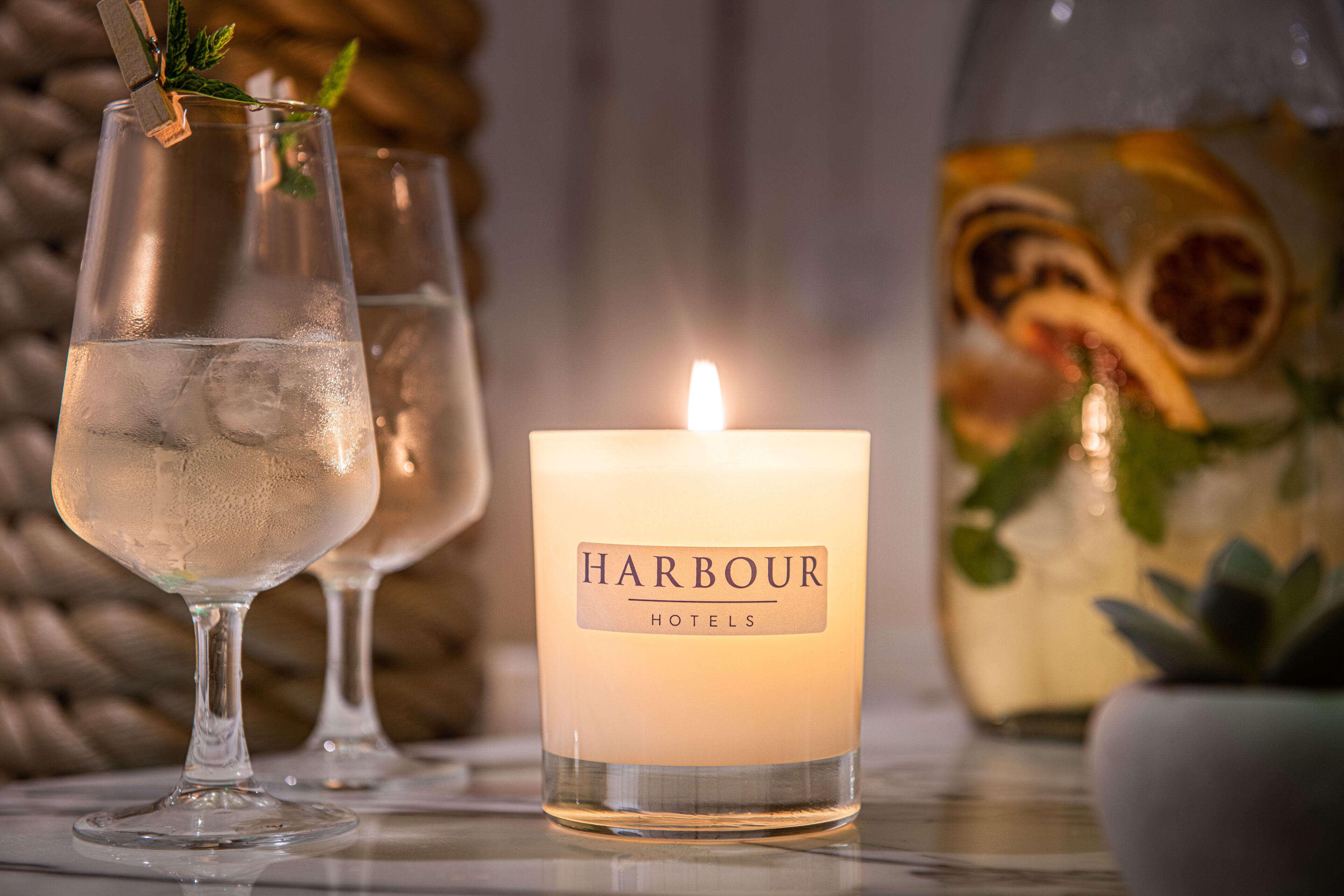 A candle labeled "Harbour Hotels" with two glasses of water and a decorative bottle with dried citrus slices.
