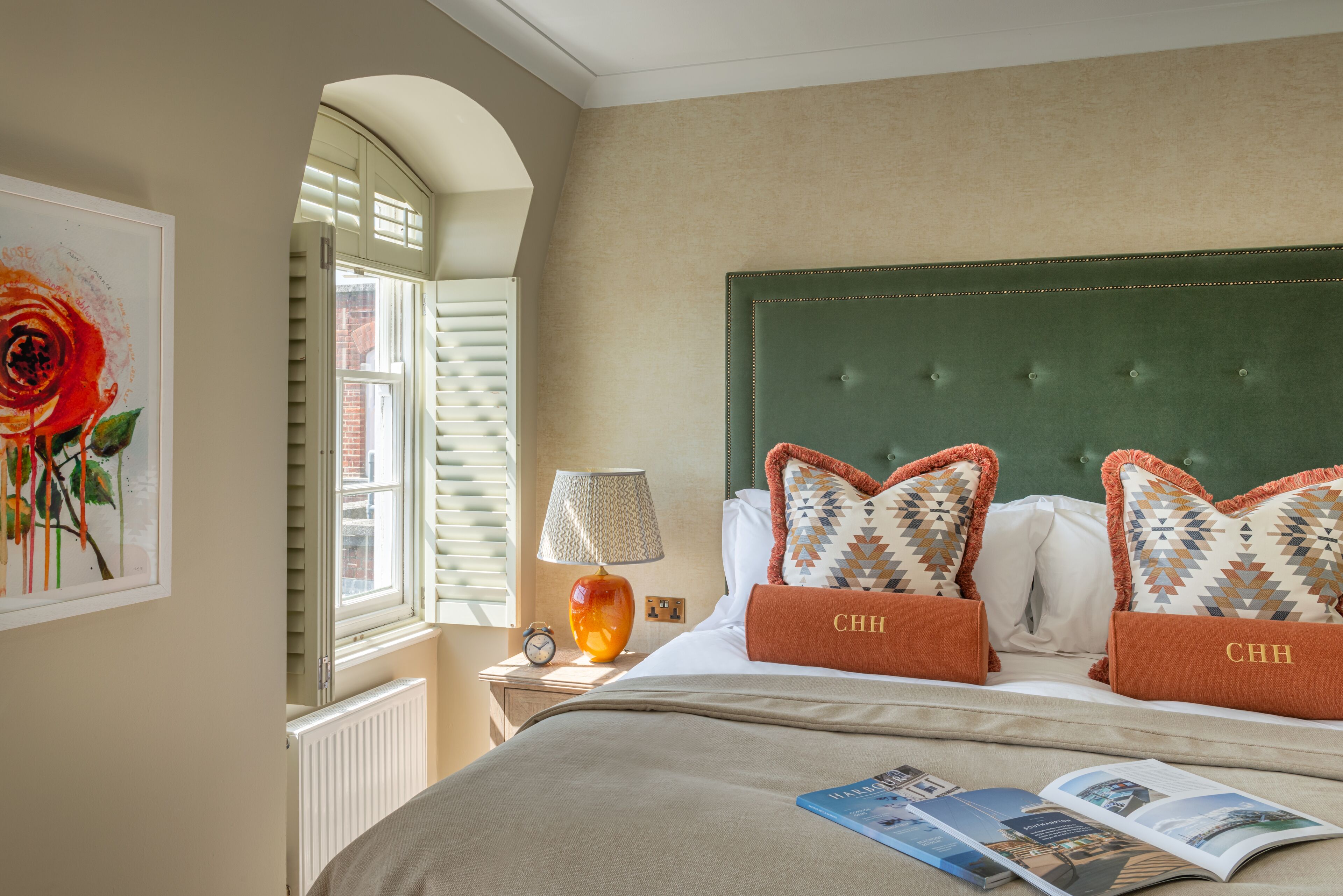 A neatly made bed with a green headboard, two pillows with 'CHH' embroidered, a lamp, a framed picture, and a magazine on the bed.