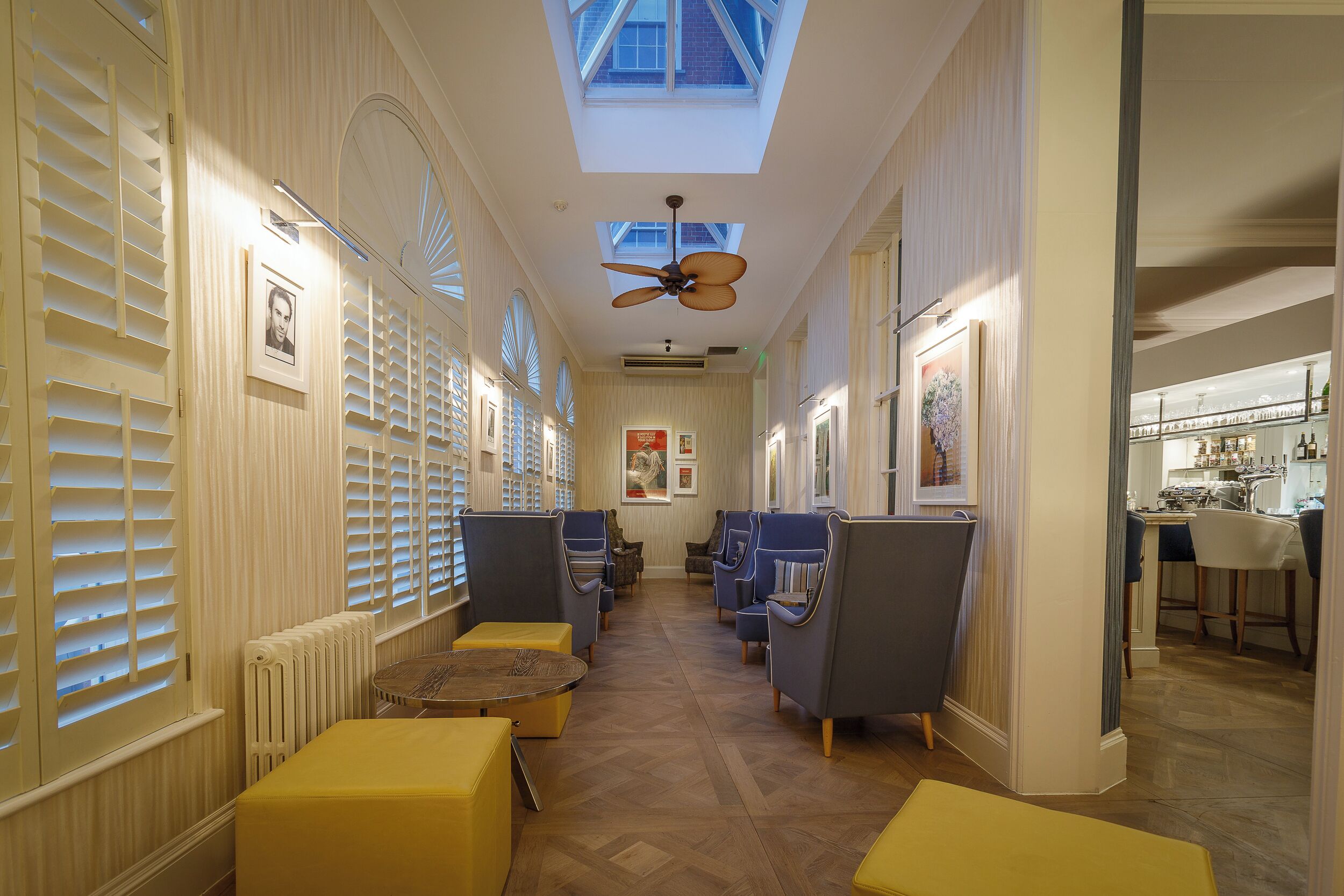 A hotel corridor with a skylight, ceiling fan, and seating area with yellow ottomans.