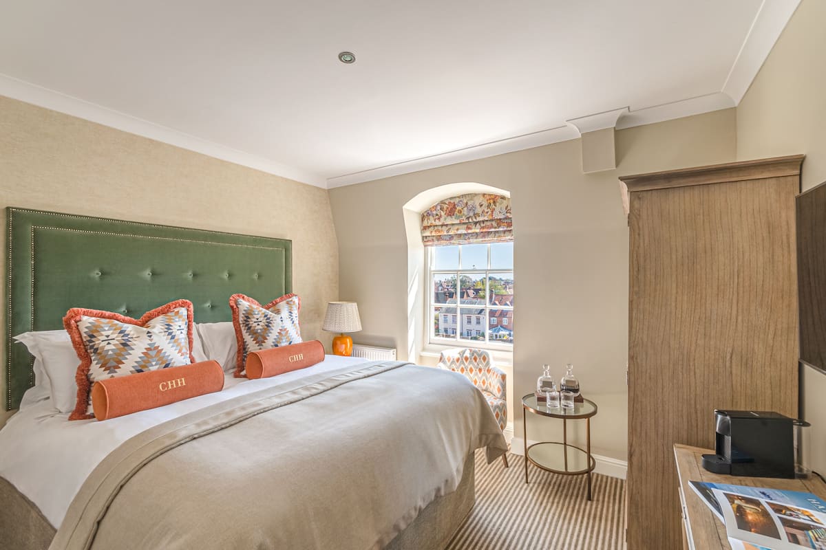 A hotel room with a large bed, a green upholstered headboard, two orange pillows, a small table with a lamp, and a view of buildings through a window.