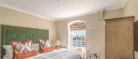 A hotel room with a large bed, a green upholstered headboard, two orange pillows, a small table with a lamp, and a view of buildings through a window.