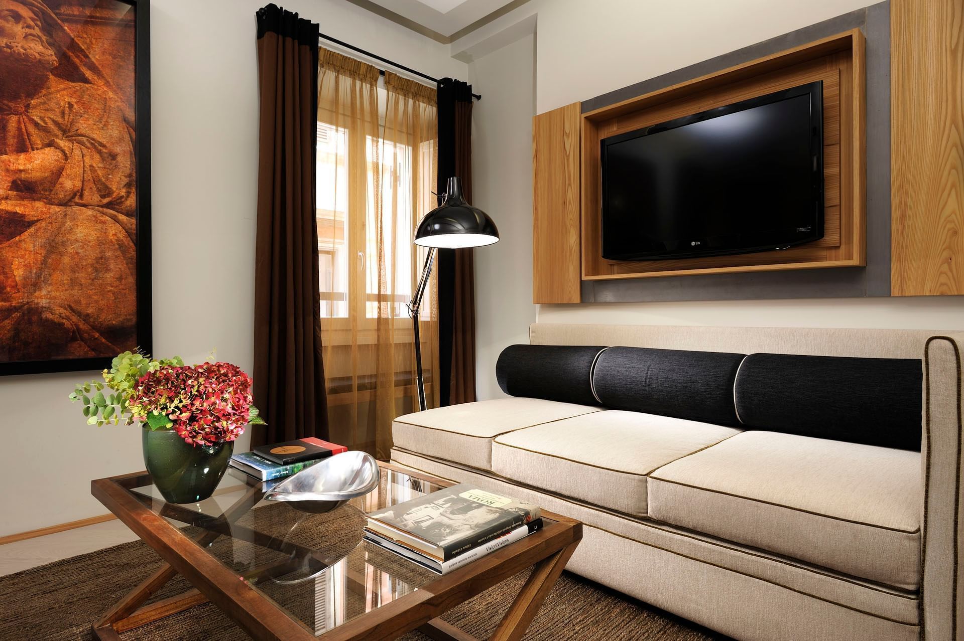 A living room with a sofa, a flat-screen TV mounted on a wooden panel, a glass coffee table with books and a vase of flowers, and a painting on the wall.
