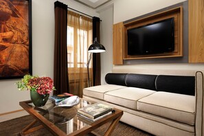 Un salon comprenant un canapé, un téléviseur à écran plat fixé sur un panneau en bois, une table basse en verre avec des livres et un vase de fleurs, ainsi qu’un tableau accroché au mur.
