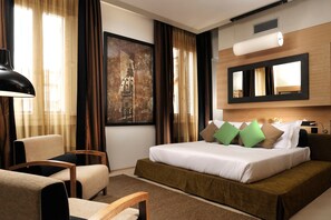 Ein Hotelzimmer mit einem Bett, einem Sessel, einem Schreibtisch, einem großen Fenster mit Vorhängen, einem Gemälde und einem Spiegel.