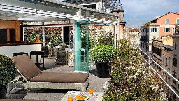 Una terraza en la azotea con un área de estar cubierta de cristal, una tumbona y una mesa con comida y bebidas.