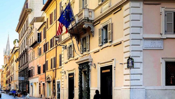 Une rangée de bâtiments historiques ornés de drapeaux européens, une rue pavée et une enseigne indiquant « VIA RIVI D’ANNO FONTANELLA ».