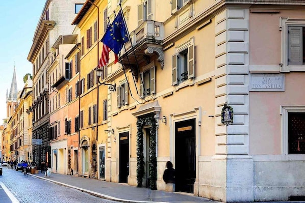 A row of historic buildings with European flags, a cobblestone street, and a sign for "VIA RIVI D'ANNO FONTANELLA". - Babuino 181 – Small Luxury Hotels of the World (Rome)