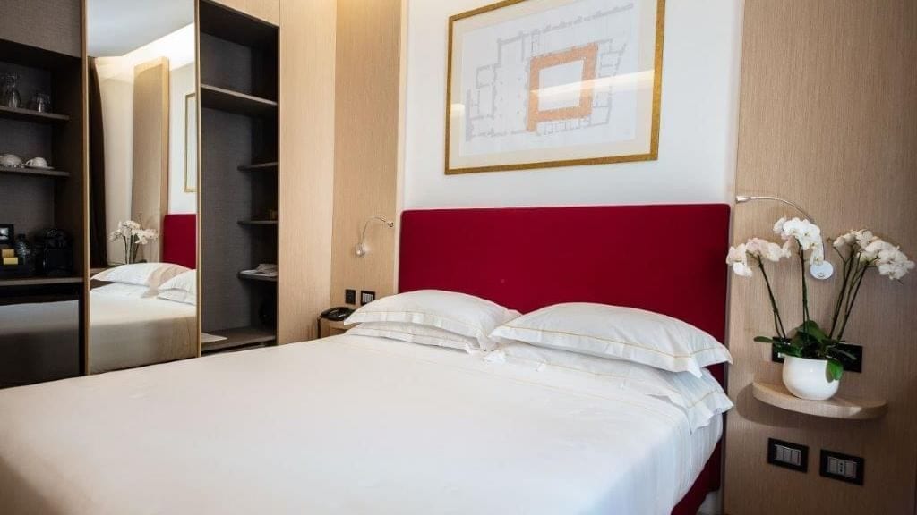 Ein Hotelzimmer mit Bett, rotem Kopfteil, Nachttisch, Spiegel, Wandpflanze und einem gerahmten Architekturplan.