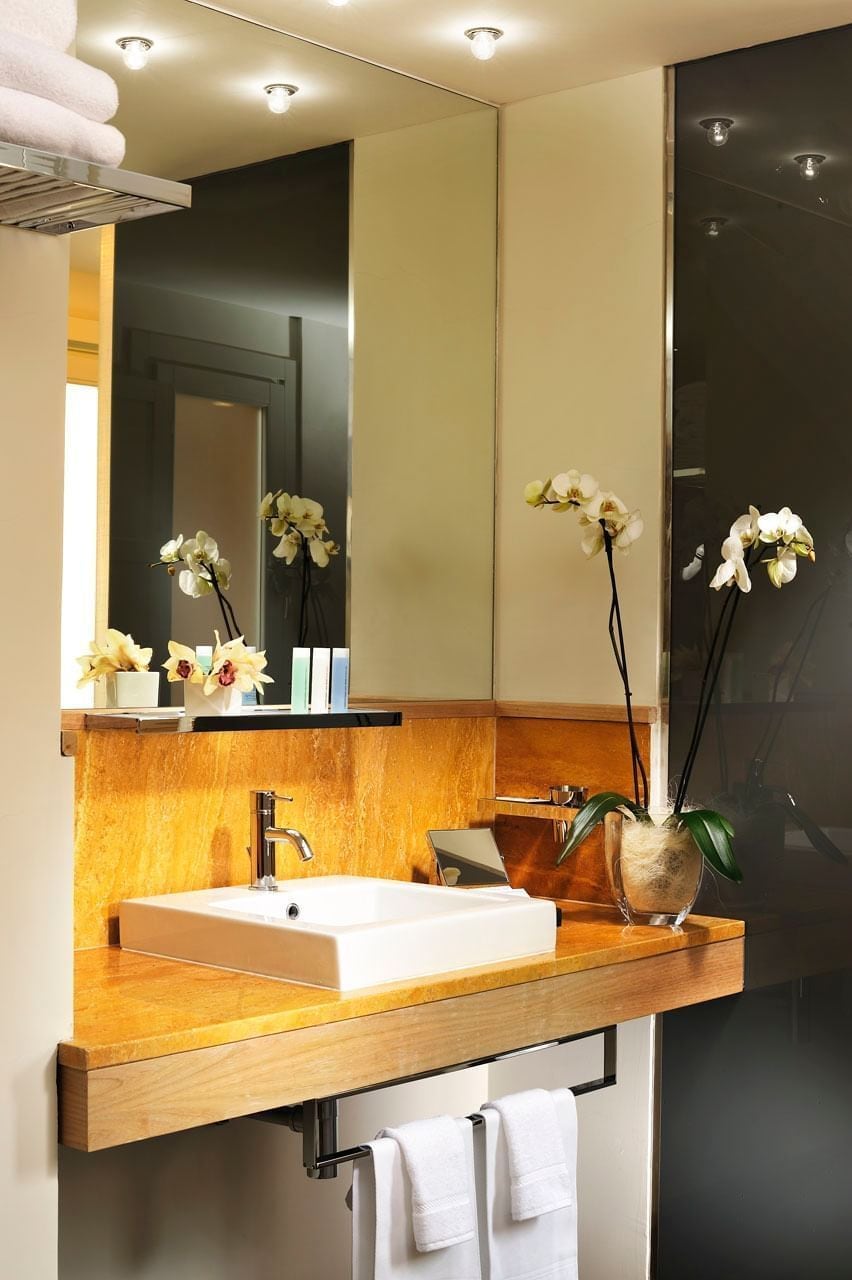 Ein modernes Badezimmer mit einem Holzlagerflächenmöbel, einem weißen Waschbecken und einem Spiegel darüber. Weiße Handtücher hängen an einem Ablagegestell unter dem Waschtisch.