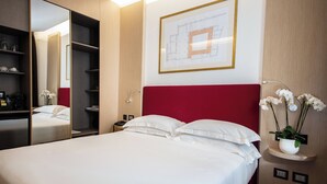 Une chambre d’hôtel avec un grand lit, une tête de lit rouge, un miroir, une table de chevet, une plante fixée au mur et un plan architectural encadré.