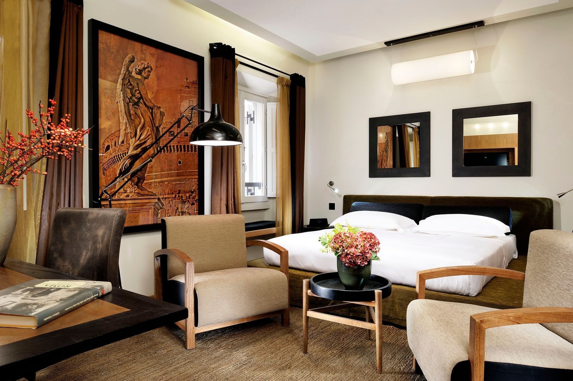 Ein Hotelzimmer mit einem Bett, zwei Stühlen, einem Schreibtisch mit Lampe, einer Vase mit Blumen und Kunstwerken an den Wänden.
