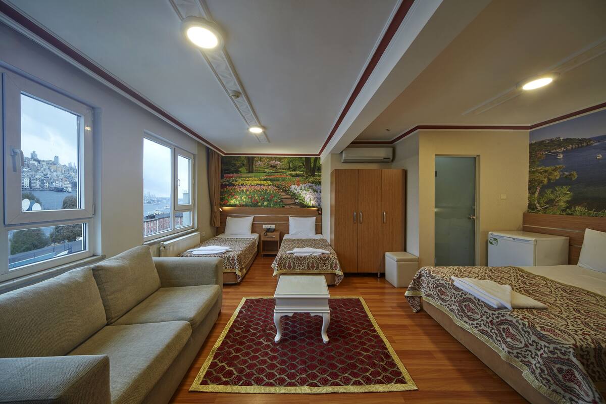 A hotel room with a city view, two beds, a sofa, a small table, and a red patterned rug.