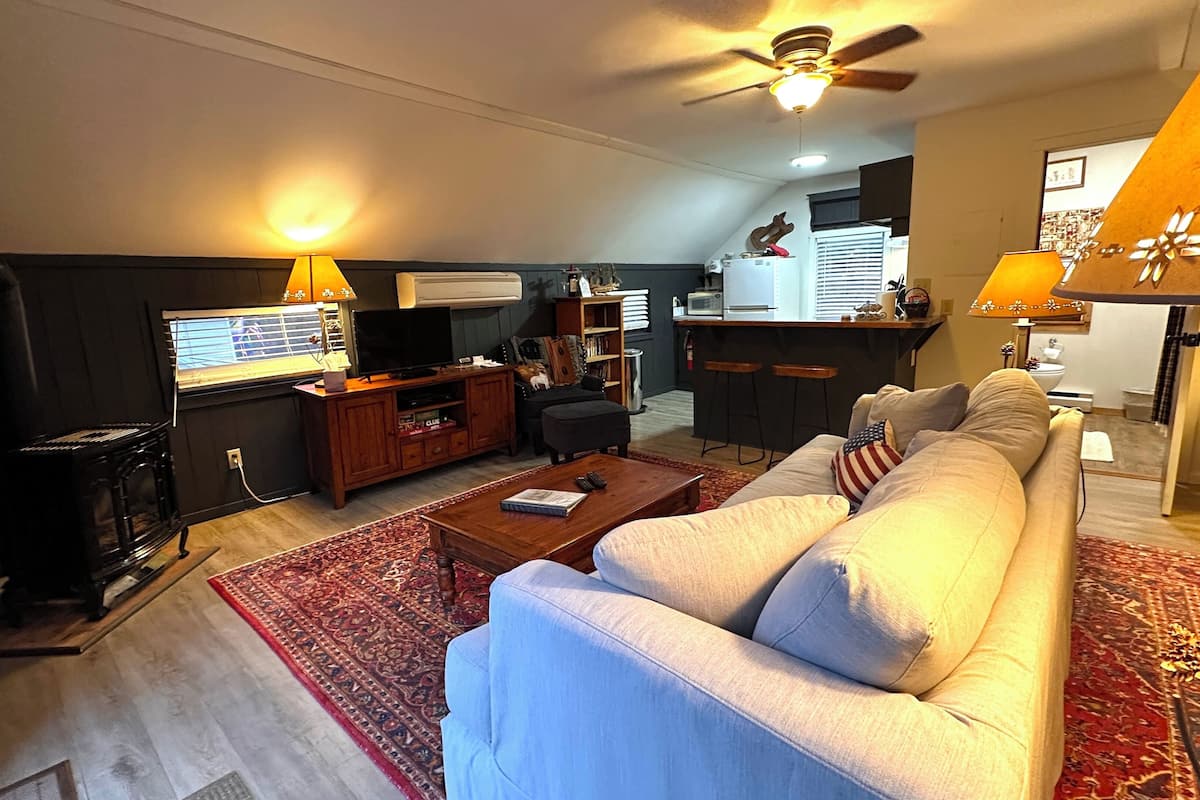 A living room with a sofa, coffee table, and a TV. There is a kitchen area in the background with a microwave and a refrigerator.