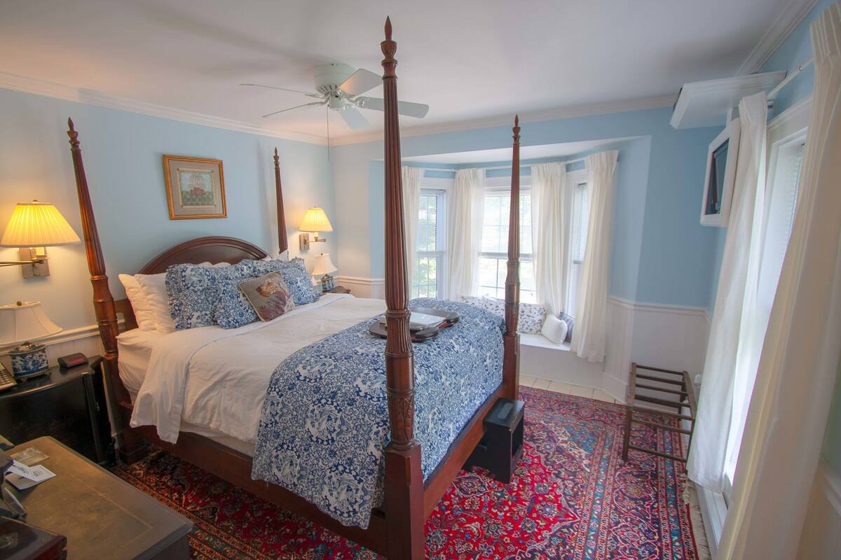 A four-poster bed with a blue and white patterned bedspread, a wooden headboard, and a red patterned rug. There is a ceiling fan, two bedside lamps, and a window with white curtains.