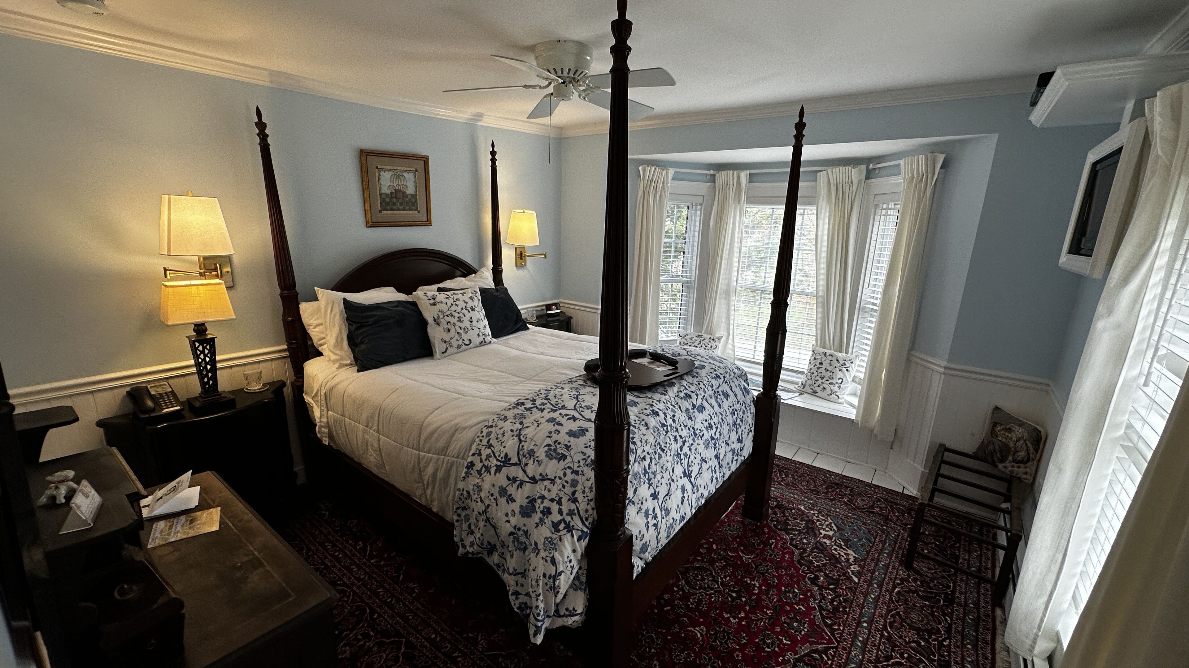 A four-poster bed with a canopy, a nightstand with a lamp, a framed picture on the wall, and a window with curtains.