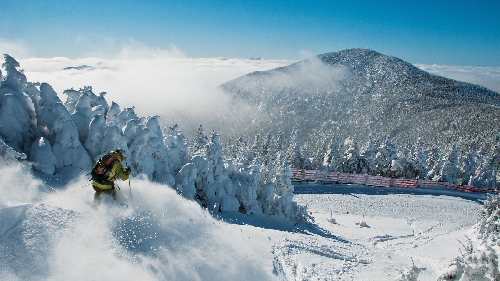 Un paysage montagneux enneigé, avec un skieur en mouvement, entouré d’arbres couverts de neige et sous un ciel dégagé.