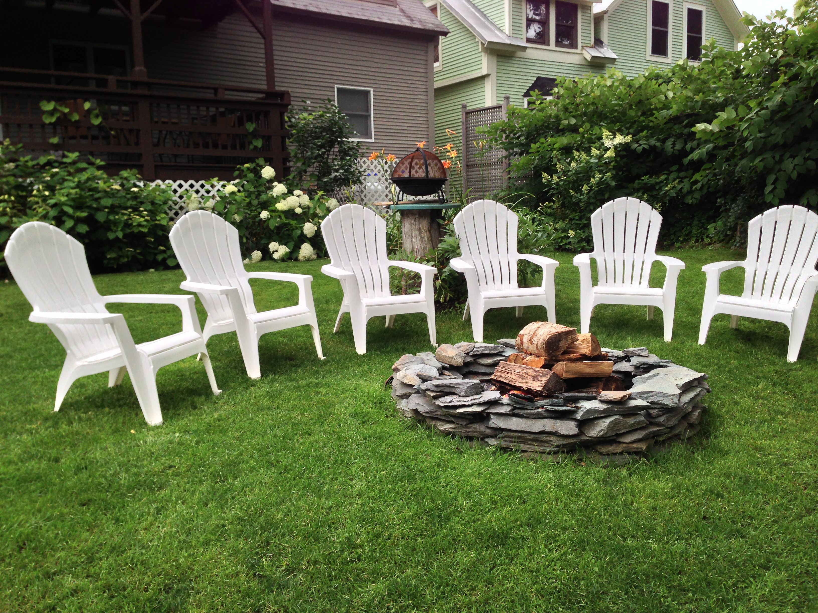 A backyard with a stone fire pit surrounded by white Adirondack chairs.