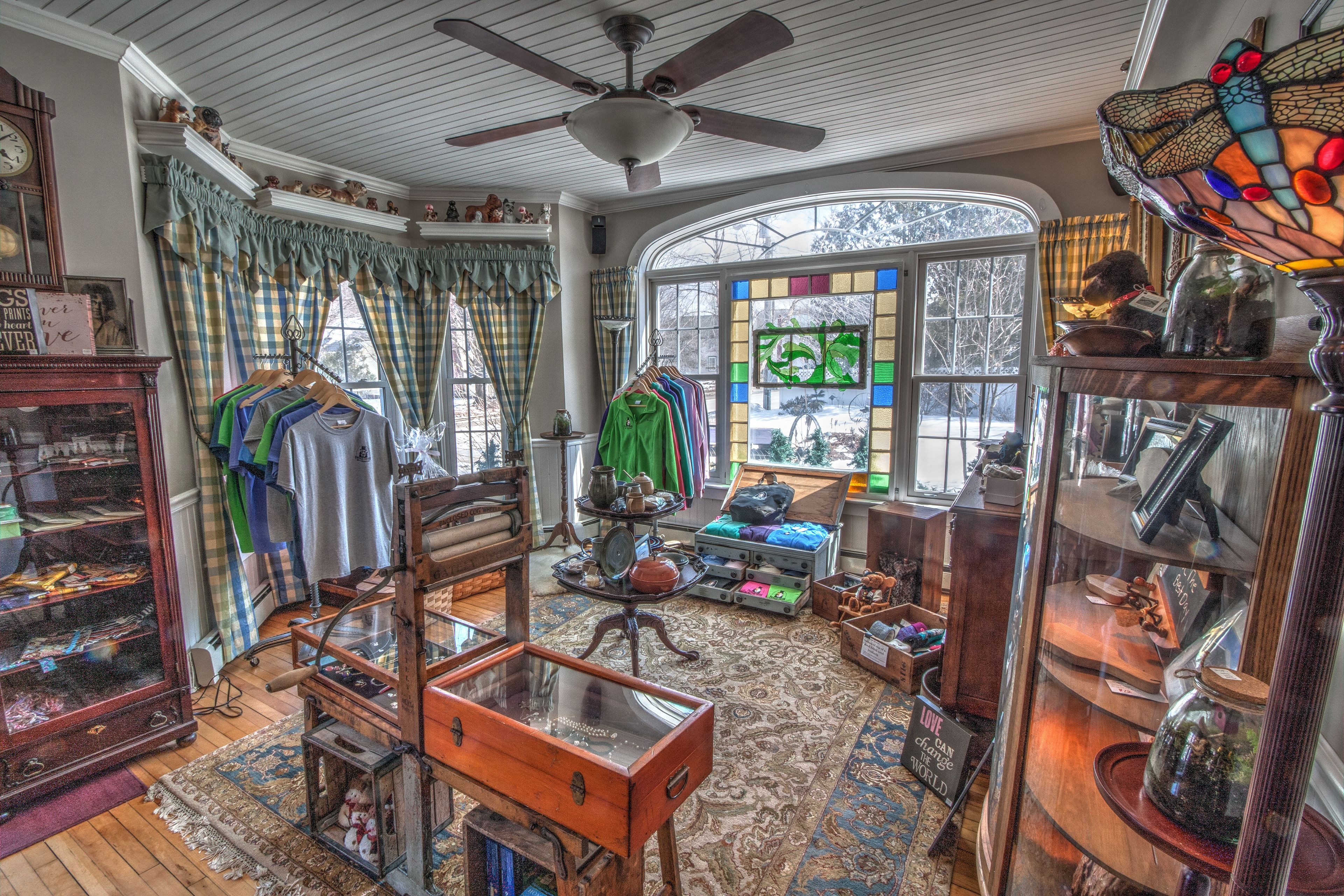 A cozy room with a large window, a ceiling fan, and various items on shelves and tables.