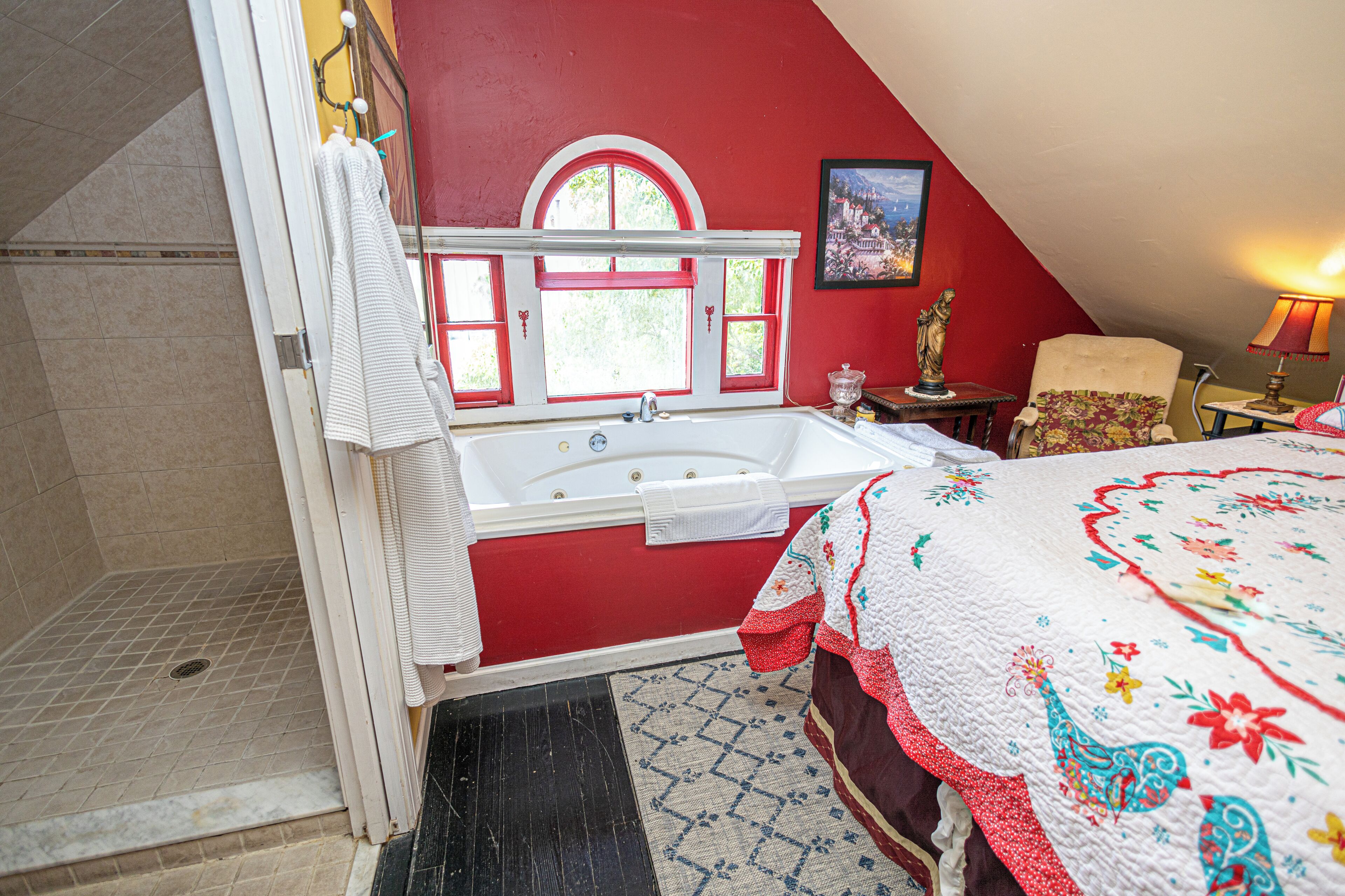A bedroom with a red wall, a large bed with a floral quilt, a window with a view of greenery, and a bathroom with a bathtub and a shower.