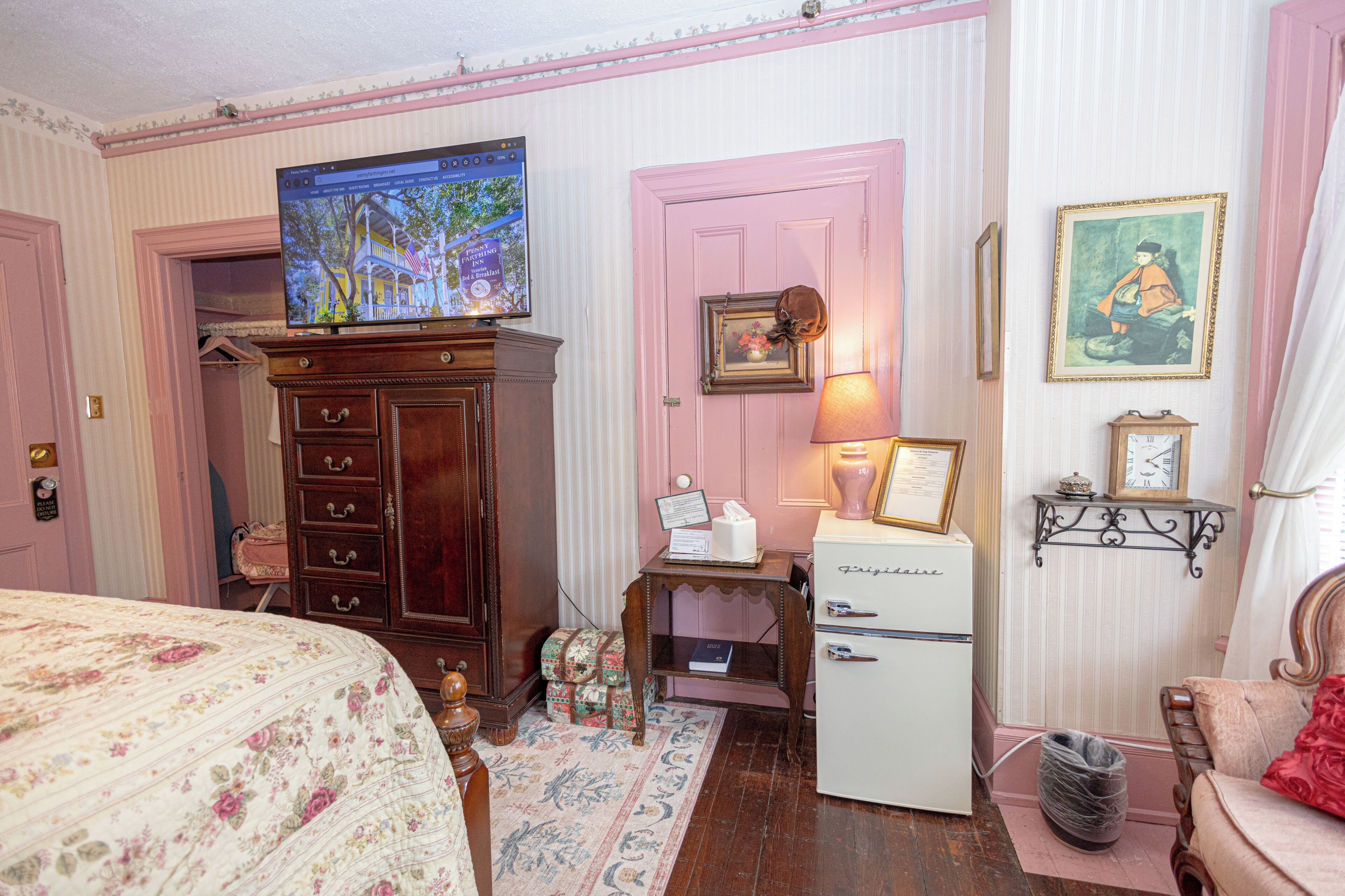 Una habitación con cama, cómoda, televisor, refrigerador y escritorio con lámpara.