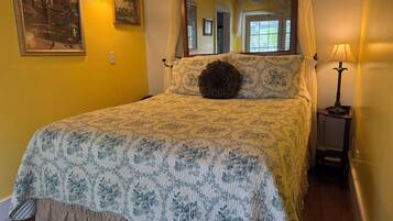 A bed with a floral bedspread, a round pillow, a nightstand with a lamp, and a painting on the wall.
