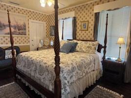 A four-poster bed with a floral bedspread, a nightstand with a lamp, and a window with blinds.