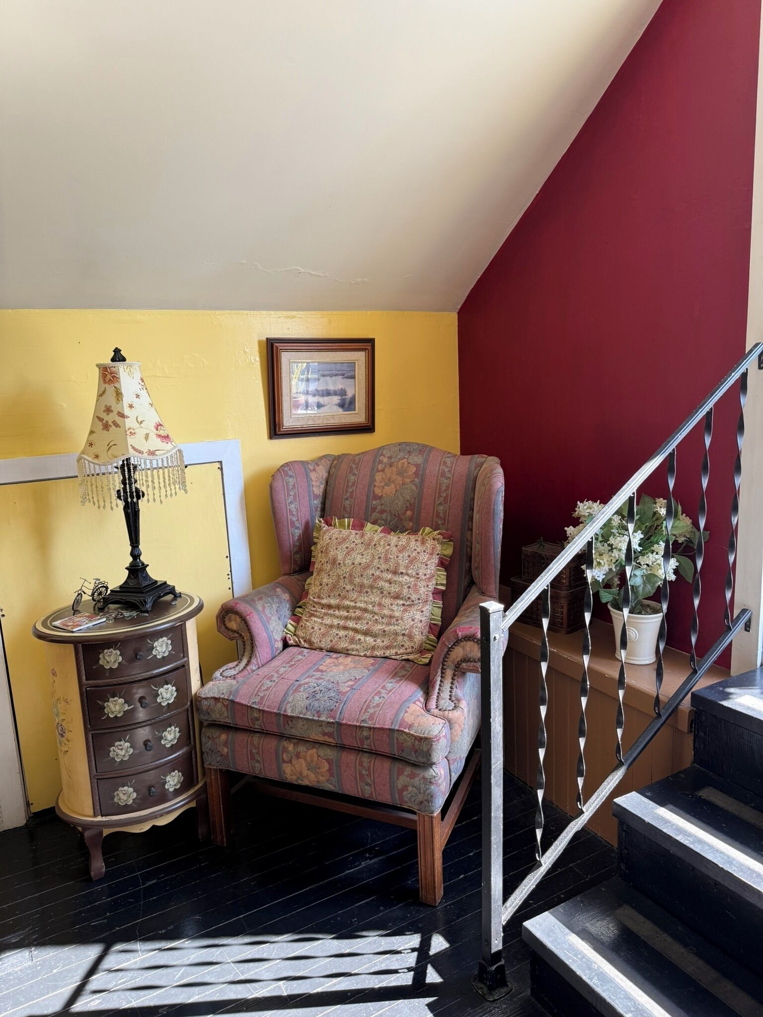 A chair with floral upholstery, a lamp with a floral shade, a framed picture on the wall, and a staircase with a railing.