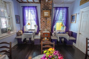 Une pièce avec un mur en briques, deux fenêtres ornées de rideaux violets, un banc en bois, une table avec une horloge et une porte.