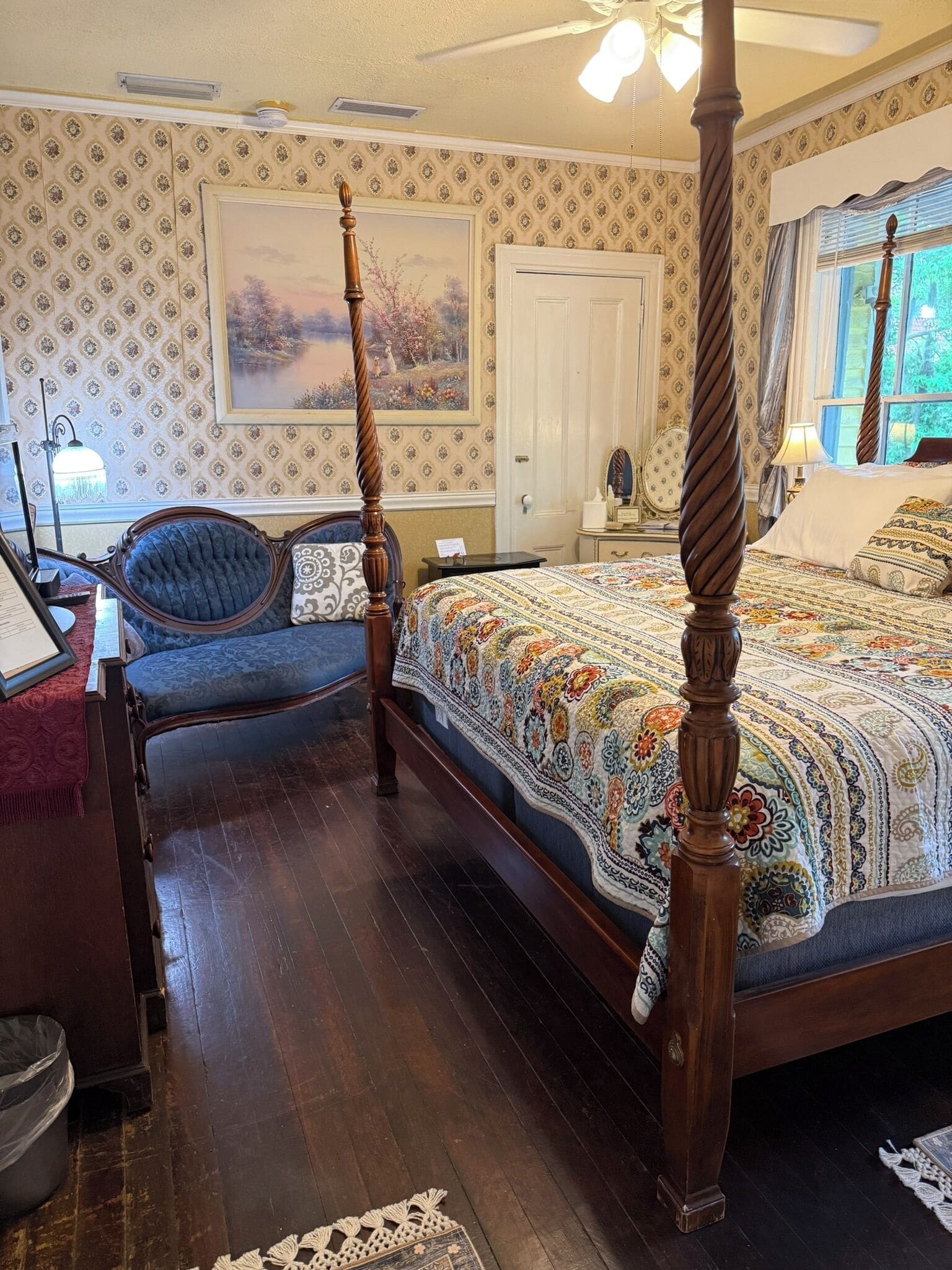 A bedroom with a four-poster bed, a chair, a desk, and a painting on the wall.