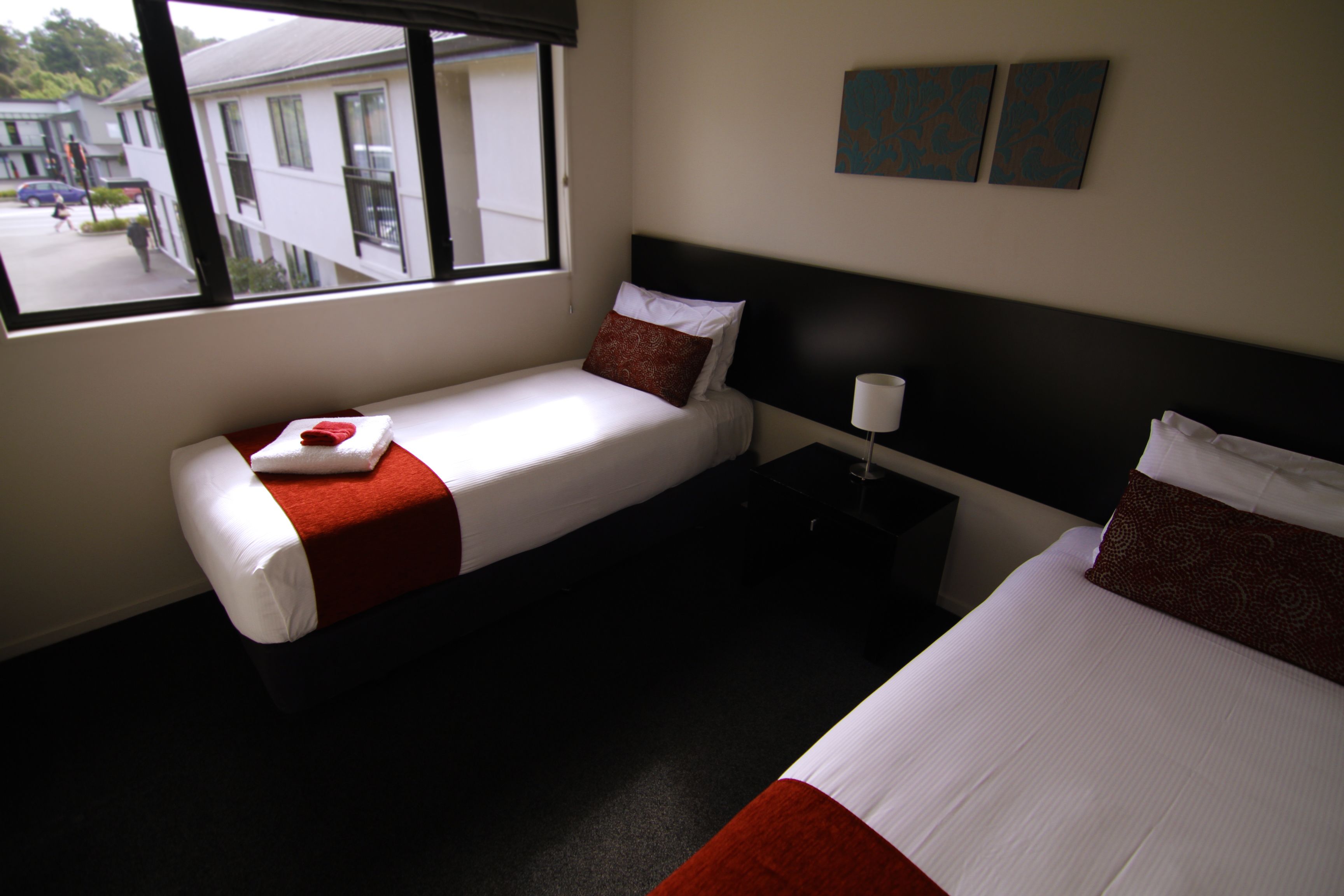 A hotel room with two beds, a window with a view of the street, and a nightstand with a lamp.