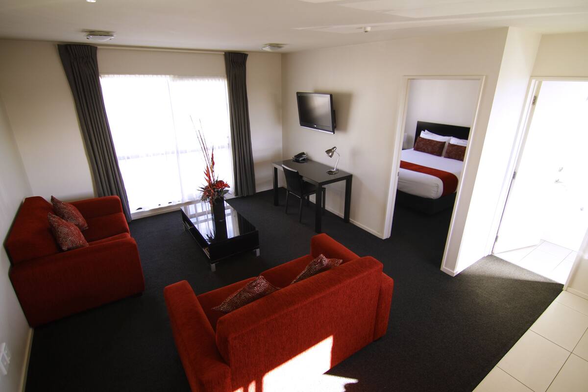 Ein modernes Hotelzimmer mit einer roten Couch, einem schwarzen Couchtisch, einem Schreibtisch mit Computer, einem Flachbildfernseher und einem Bett mit weißer und brauner Bettwäsche.