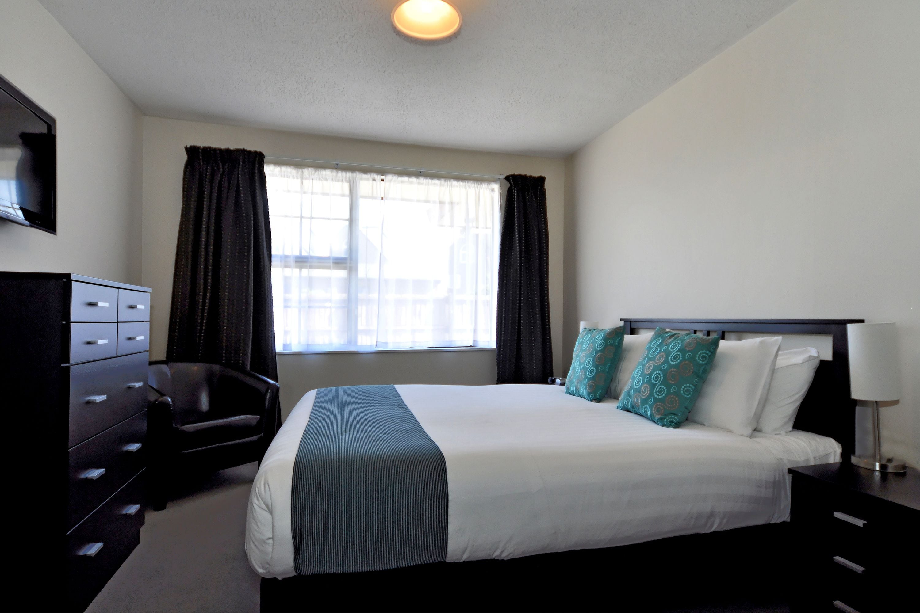 A hotel room with a bed, a dresser, a chair, a window with curtains, and a lamp.