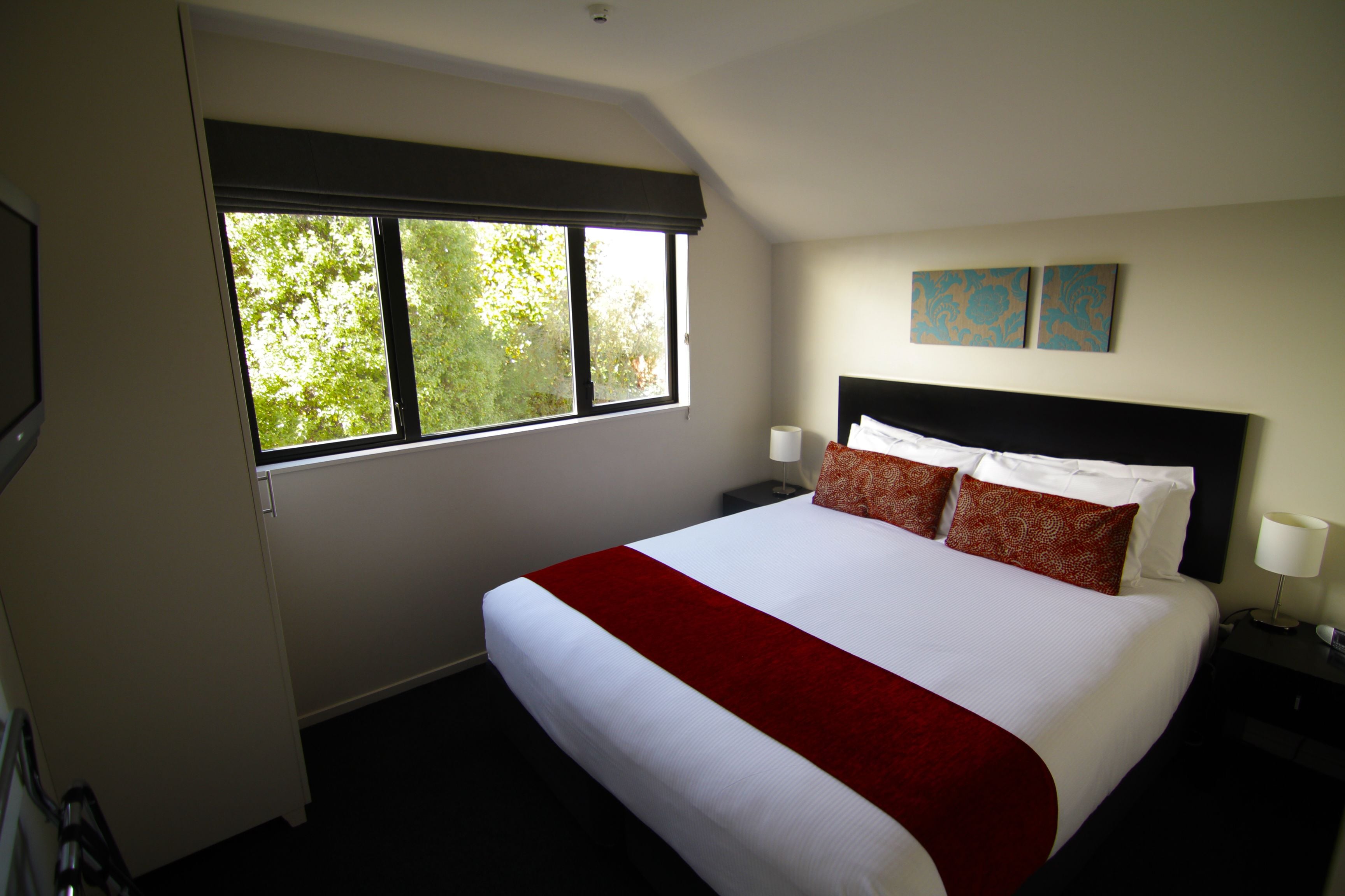 A hotel room with a large bed, two bedside tables, a window with a view of trees, and two wall-mounted artworks.