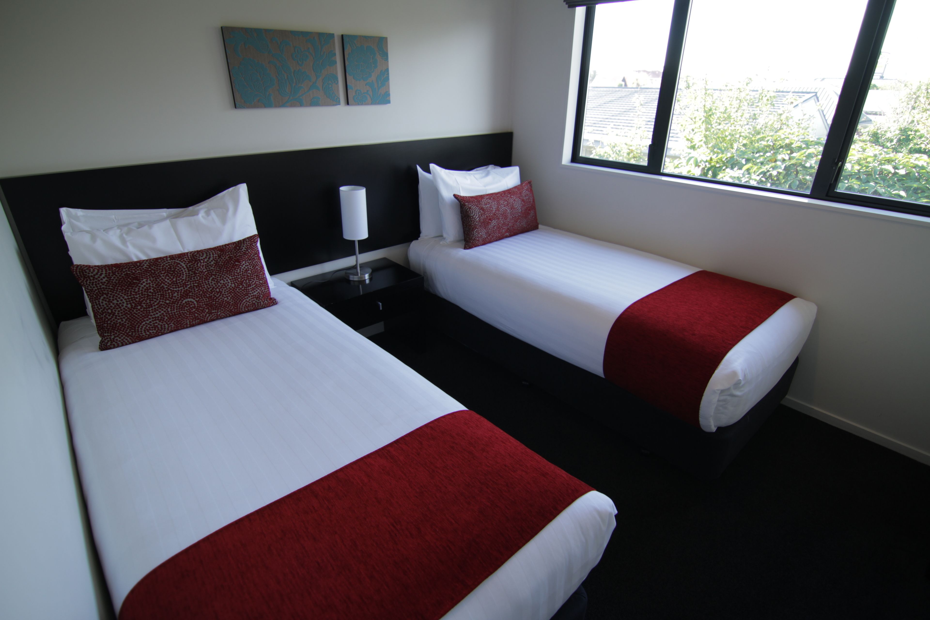 Habitación de hotel con dos camas, cada una con ropa blanca y detalles rojos, una mesita de noche con lámpara y una ventana con vistas a la vegetación.