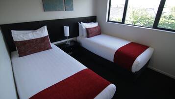 Habitación de hotel con dos camas, cada una con ropa blanca y detalles rojos, una mesita de noche con lámpara y una ventana con vistas a la vegetación.