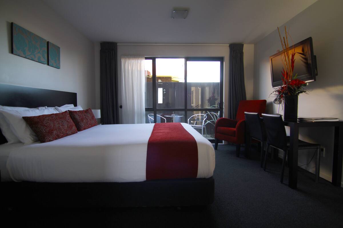 Ein Hotelzimmer mit einem großen Bett, einem Schreibtisch, einem Sessel, einem Fernseher und einem Fenster mit Vorhängen.