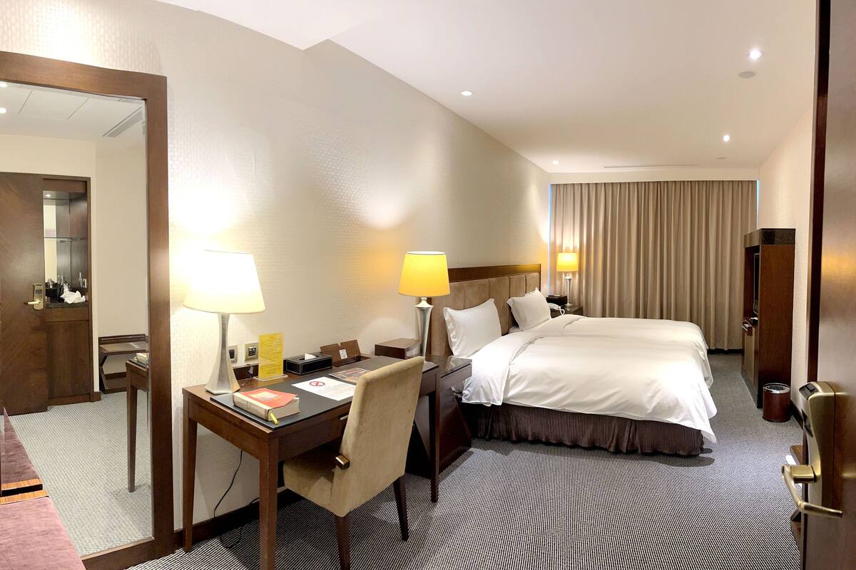 A hotel room with a large bed, two bedside tables with lamps, a desk with a chair, a mirror, and a wardrobe.