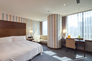 Quarto de hotel com uma cama grande, um sofá, uma mesa de escritório com uma cadeira e uma janela ampla com cortinas.