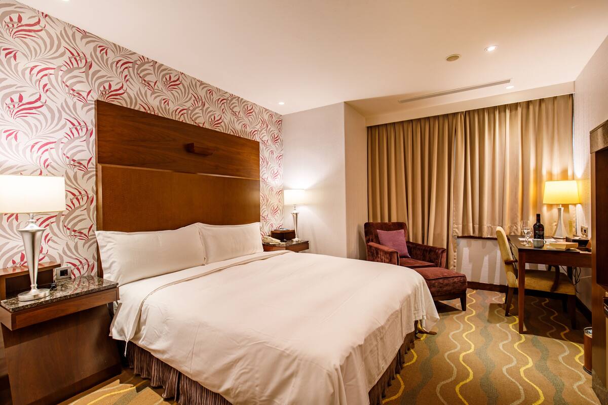 A hotel room with a large bed, a desk with a chair, a small table with a lamp, and a window with curtains.