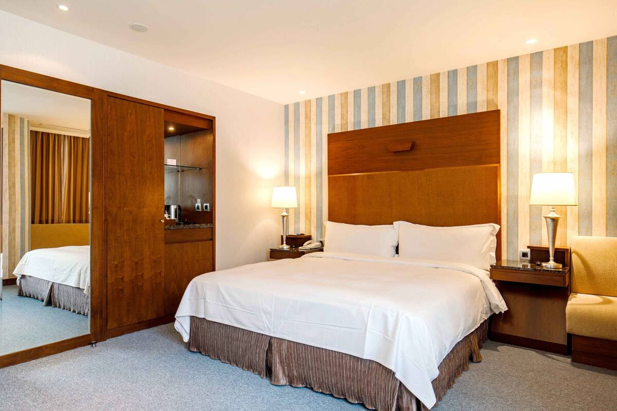 A hotel room with a large bed, wooden headboard, two bedside tables with lamps, a mirror, and a striped wall.