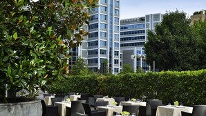 An outdoor dining area with tables and chairs, surrounded by greenery and a modern building in the background. - UNA Hotels The One Milano (San Donato Milanese)