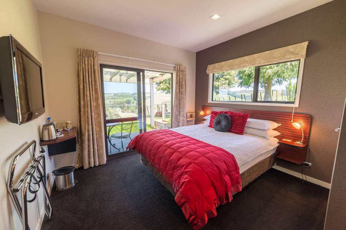 A bedroom with a large bed, a window with a view, a bedside table with a lamp, and a small table with a kettle and a pot.